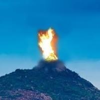 Close view of the summit of Arunachala hill against a blue sky with a large flame rising from the peak.