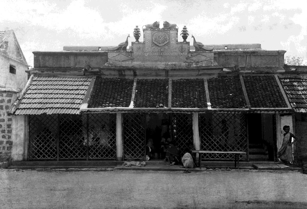 Black-and-white photograph of the front of a single-storey house with tiled roofs and lattice screens; a signboard on the parapet reads SRI SUNDARAMANDIRAM with Tamil above; a woman stands at the right and a dog lies by the steps.