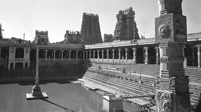 Black-and-white view across the stepped temple tank of the Meenakshi Sundareswarar Temple, Madurai, with pillared colonnades around the water, a lamp post in the tank, a carved stone pillar at right and gopurams rising behind. Whole picture.