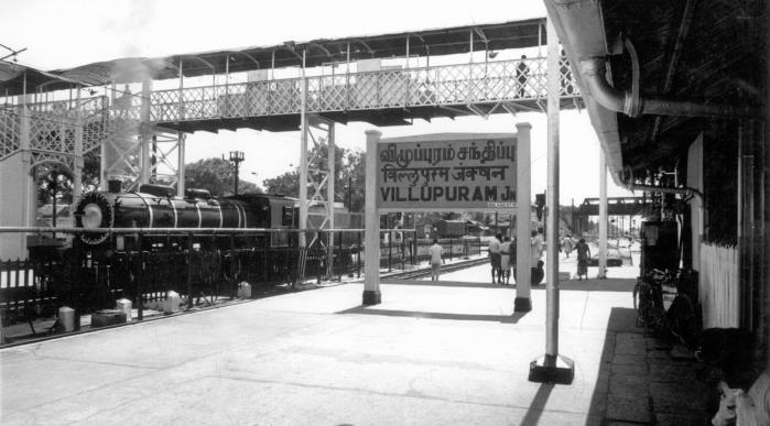 Black-and-white railway platform with a steam locomotive at left, a lattice footbridge overhead and a station name board in Tamil, Hindi and English reading VILLUPURAM Jn. Whole picture.