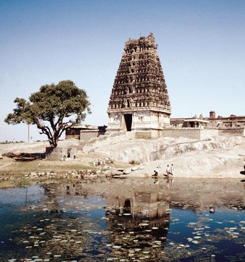 Colour photograph of a temple gopuram standing on a large bare rock beside a tree, reflected in a pond in the foreground. Whole picture.
