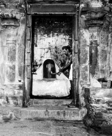 Black-and-white view through a stone doorway into a dark sanctum where a priest tends a Siva lingam draped in white cloth. Whole picture.