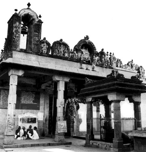 Black-and-white photograph of a small temple entrance with a bell arch and carved figures on the parapet, pillared porch and iron-railed shrine, with several people seated on the ground at left. Whole picture.