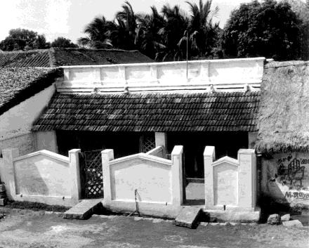 Black-and-white photograph of a single-storey house with a tiled roof behind a low white wall with gateposts, coconut palms behind. Whole picture.