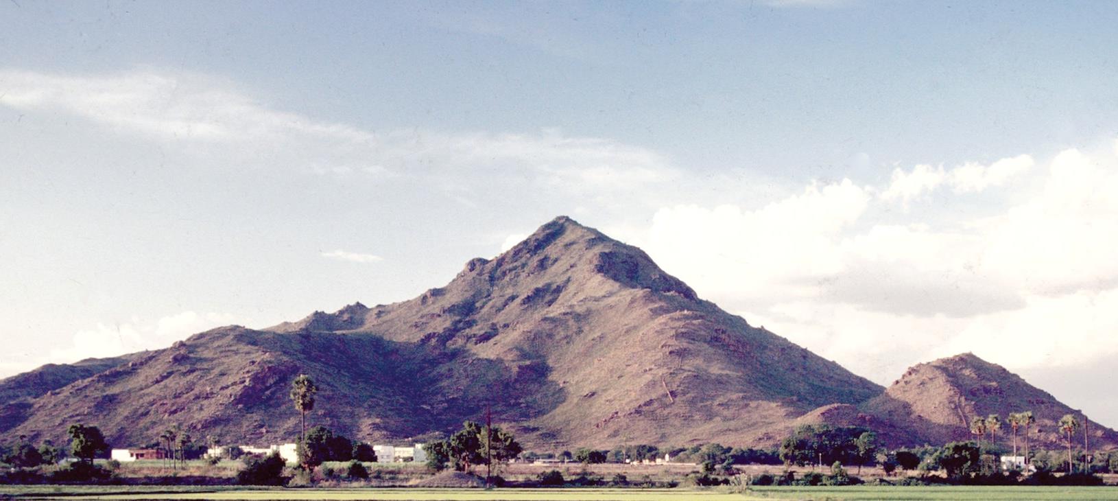Colour panorama of Arunachala hill under a pale blue sky with clouds, with trees, palms, white buildings and green fields at its foot. Whole picture.