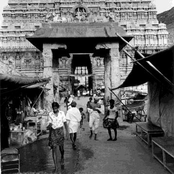 Black-and-white street scene of people walking on a wet lane lined with stalls and awnings in front of a stone mantapam, with a carved gopuram behind. Whole picture.