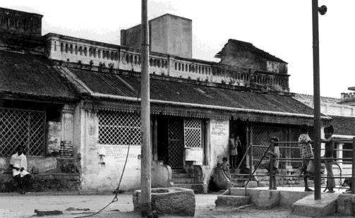 Black-and-white photograph of a row of old houses with tiled verandas and lattice windows along a street, with children at a railing and a man seated at left. Whole picture.