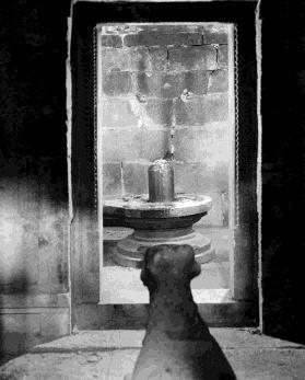 A Siva lingam on its pedestal seen through a dark doorway of a small shrine.