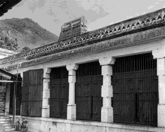 A long pillared building with barred wooden doors between the pillars, a hill in the background; a bicycle at the left.