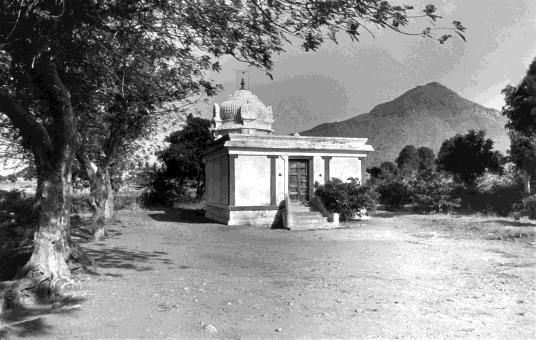 A small white temple with a domed roof in a clearing under trees, a conical hill behind.