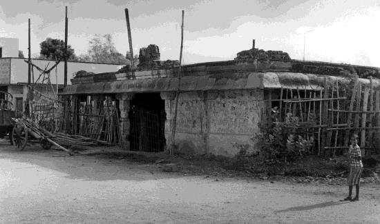A low, weathered stone temple by a dirt road, with scaffolding poles and a child standing at the right.