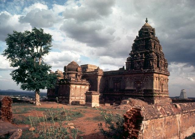 Colour photograph of a stone temple with a tall tower on a hilltop under a cloudy sky, a tree at the left.