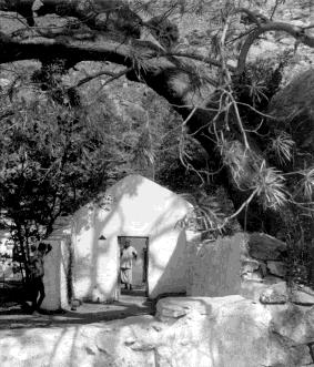 A small whitewashed structure built against rock under an overhanging tree, a figure in its doorway.