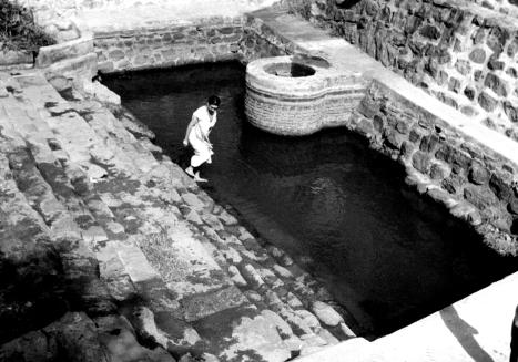 A stone-walled water tank with steps, a round well-like structure at the back, a person stepping into the water.