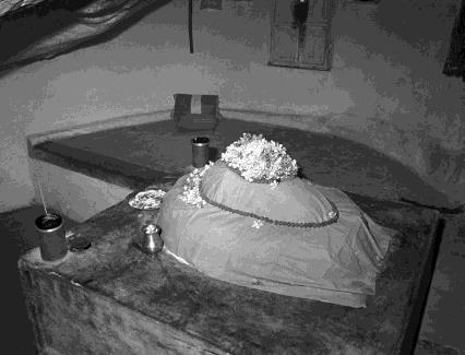 A mound-shaped linga covered in cloth and flowers on a platform inside a cave, with small vessels beside it.