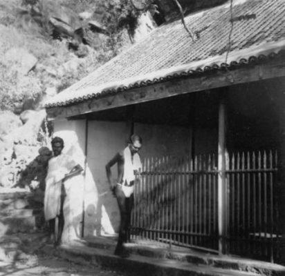 The front of a building with a tiled roof and iron railings, two men standing by the entrance.