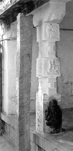 Black-and-white photograph of a carved stone pillar and granite post inside an old stone mantapam, with a dark stain at the pillar base. Whole picture.