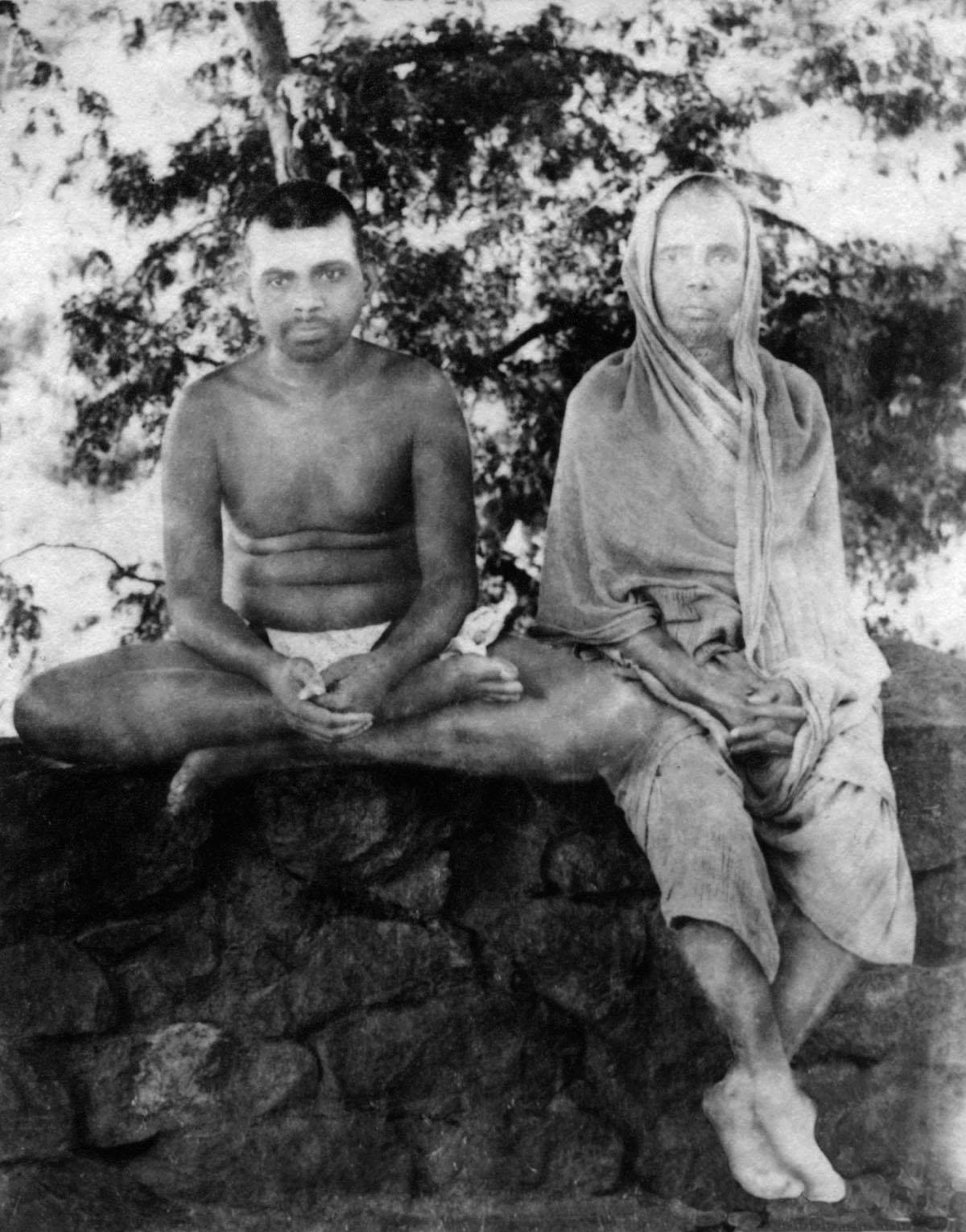 Black-and-white photograph of the young Sri Ramana Maharshi, bare-chested in a loincloth, seated cross-legged on a rough stone wall beside his mother Alagammal, who sits with her head covered by a cloth; trees behind. Whole picture.