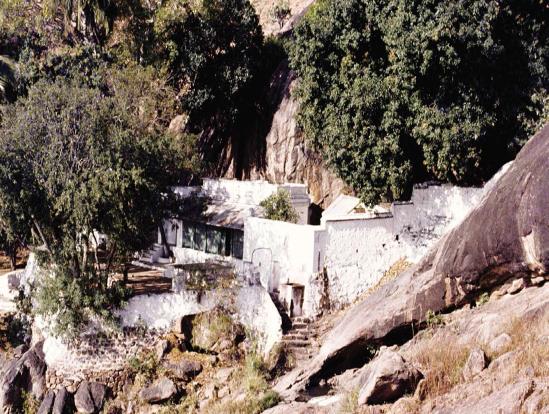 Colour photograph of Skandashram: white-washed buildings and walls set among trees against a large rock face on the hillside, with steps leading up. Whole picture.