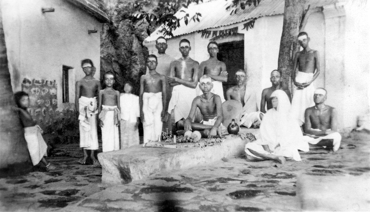 Black-and-white group photograph outside Skandashram: Sri Ramana Maharshi seated on a low stone platform, his mother in white seated nearby, and a group of men and boys standing and sitting around them in front of the whitewashed buildings. Whole picture.