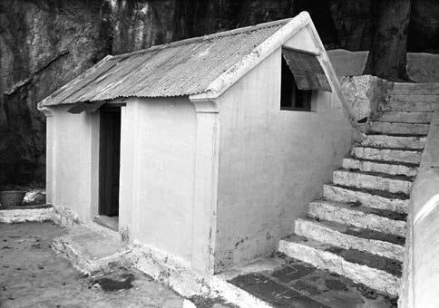 Black-and-white photograph of a small whitewashed building with a corrugated sloping roof and a flight of stone steps beside it against a rock face. Whole picture.