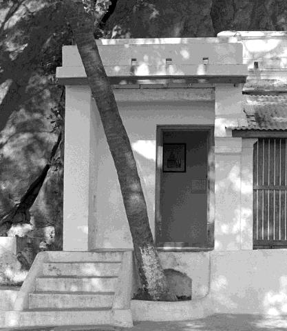 Black-and-white photograph of a small whitewashed room with a doorway, barred window and short flight of steps, with a leaning palm trunk in front. Whole picture.