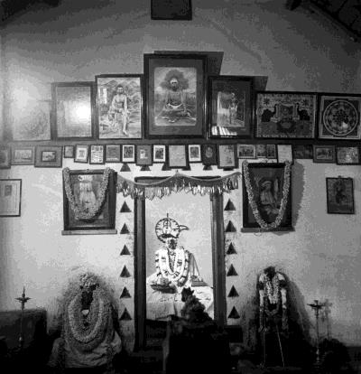 Black-and-white photograph of the interior of the early shrine: a wall hung with framed pictures above a garlanded doorway lined with triangular lamps, with garlanded figures on either side. Whole picture.