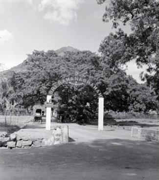 Black-and-white photograph of an arched entrance gate lettered SRI RAMANASRAMAM on a road, with large trees and the hill behind. Uncaptioned. Whole picture.
