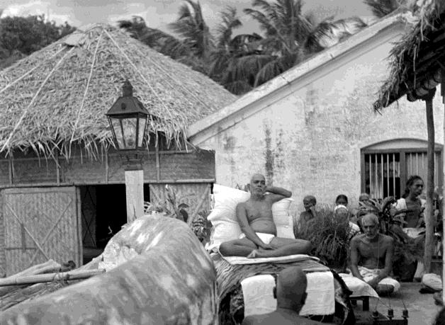 Black-and-white photograph of Sri Ramana Maharshi reclining on a couch with bolsters outdoors beside a thatched structure and the old hall, with devotees seated around him and a lantern in the foreground. Whole picture.