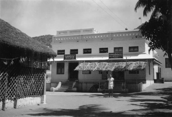 Black-and-white photograph of a two-storey white building with a thatched awning and a sign reading OFFICE; a thatched hut with lattice fence on the left and a person standing by the steps. Whole picture.