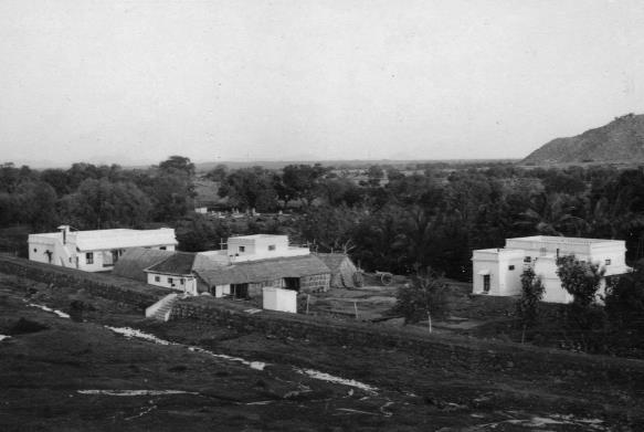 Black-and-white elevated view of several white flat-roofed buildings and tiled sheds among trees, with a hill at the upper right. Whole picture.