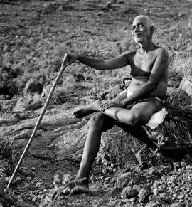 Black-and-white photograph of Sri Ramana Maharshi seated on a rock on a hillside, holding a walking staff, smiling. Whole picture; uncaptioned.