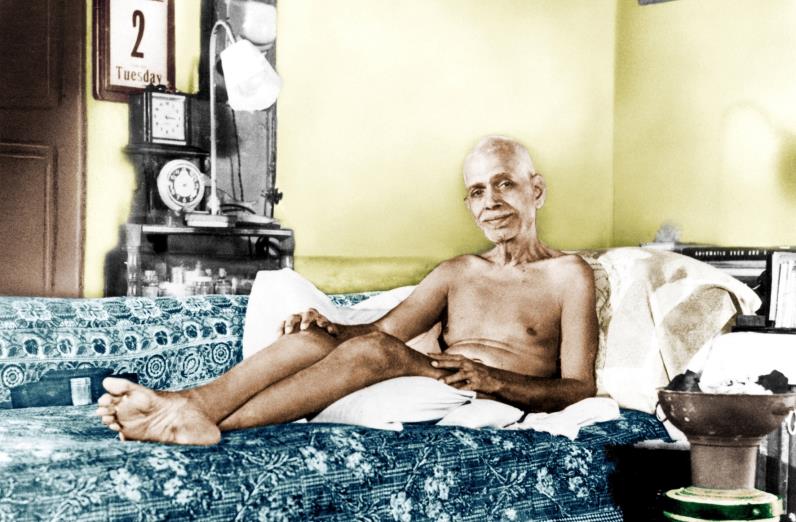 Hand-coloured photograph of Sri Ramana Maharshi reclining on a couch with a blue patterned cover, a calendar showing '2 Tuesday', clocks and a lamp behind him and a brass vessel at right. Whole picture.