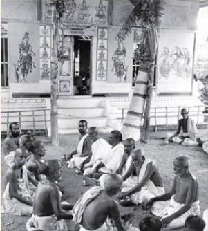 Black-and-white photograph of rows of bare-chested priests seated on the ground performing a ritual in front of a temple doorway flanked by painted figures and decorated posts. Whole picture.