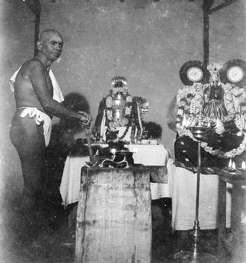 Black-and-white photograph of a man with a cloth over his shoulder standing at a shrine with garlanded images and a tall oil lamp. Whole picture.