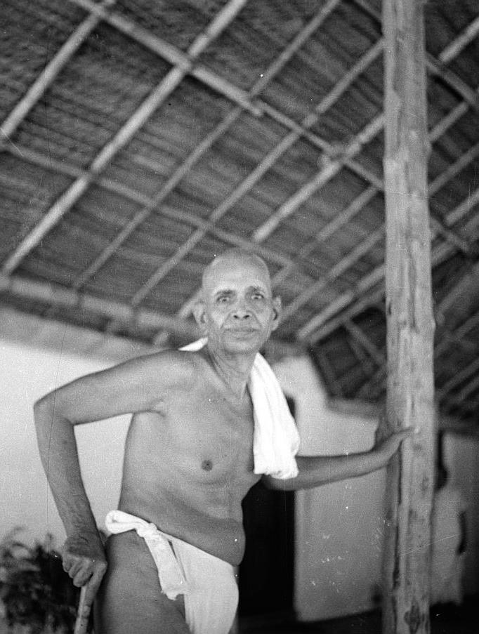 Black-and-white photograph of Sri Ramana Maharshi standing under a thatched roof, one hand on a wooden post and the other on a staff, a towel over his shoulder. Whole picture; uncaptioned.