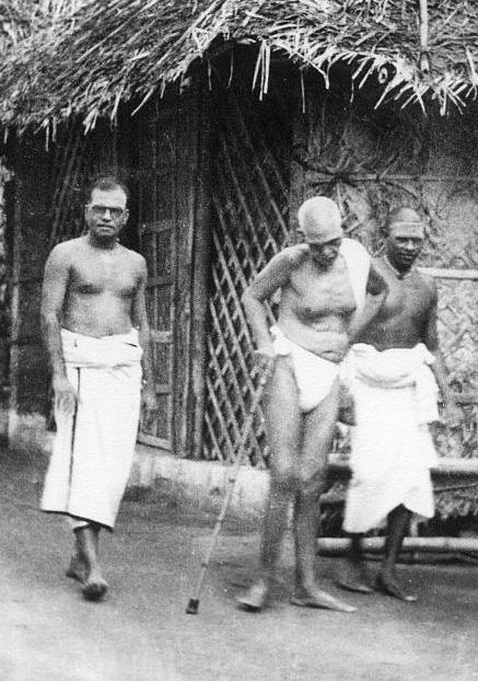 Black-and-white photograph of Sri Ramana Maharshi walking with a staff in front of a thatched hut with lattice work, with two men in white dhotis beside him. Whole picture; uncaptioned.