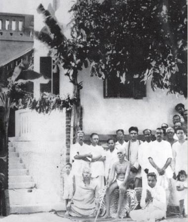 Black-and-white photograph of Sri Bhagavan seated on a tiger skin in front of a whitewashed building with steps, a large group of men and children standing around Him.