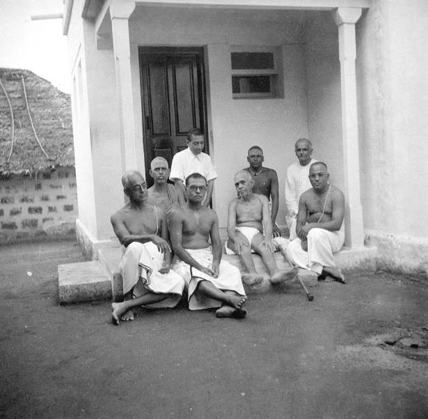Black-and-white photograph of Sri Bhagavan seated with a group of men on the steps of a small pillared building beside a thatched roof.