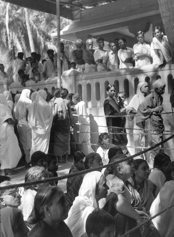 Black-and-white photograph of a crowd of devotees, many women in saris, waiting behind ropes in a queue outside a building with a balustrade.