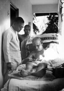 Black-and-white photograph of Sri Bhagavan reclining on a couch while two attendants stand beside Him.