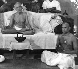 Black-and-white photograph of Sri Bhagavan seated cross-legged on a couch, with a kettle on a small table in front and a man seated on the floor to the right.