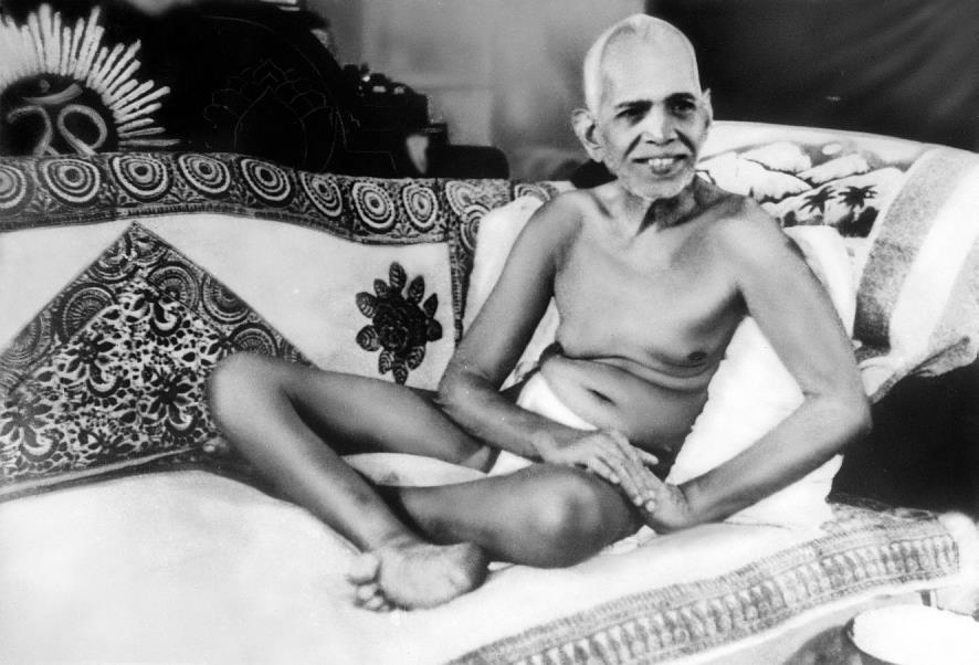 Black-and-white photograph of Sri Bhagavan smiling, seated on a couch with patterned covers, one knee raised, an Om emblem on the backrest behind.