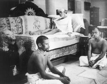 Black-and-white photograph of Sri Bhagavan reclining on a couch in a hall, with two men seated on the floor in front.