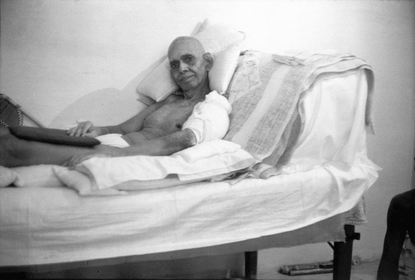 Black-and-white photograph of Sri Bhagavan reclining on a bed against pillows, His left arm bandaged, a patterned cloth folded beside Him.
