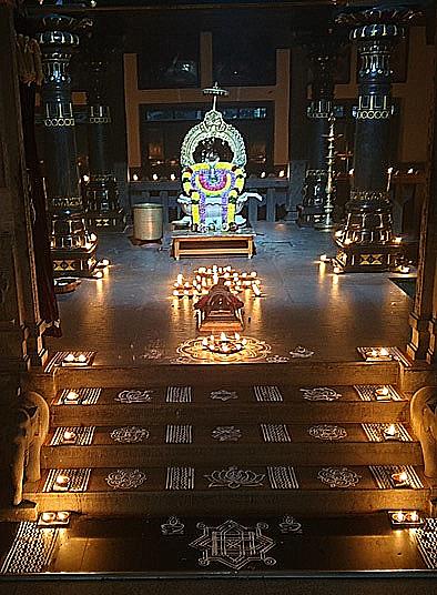 Colour photograph at night of a shrine with a garlanded samadhi and arch, oil lamps on the floor and kolam designs on the steps in front.