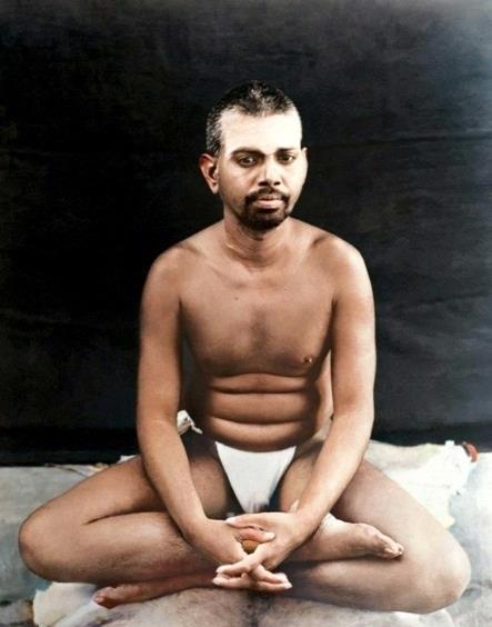 colour photo of the young Sri Bhagavan seated cross-legged, hands clasped, dark background