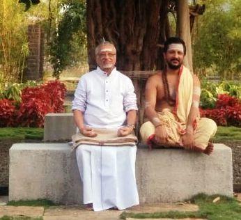 photo of two men seated on a stone bench in a garden, one in white, one bare-chested with a dhoti