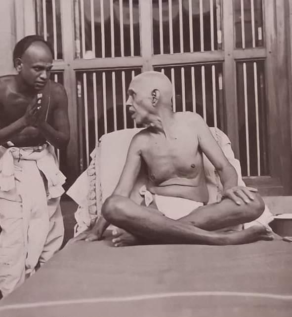 sepia photo: Sri Bhagavan seated cross-legged on a couch, a man with folded hands standing at left