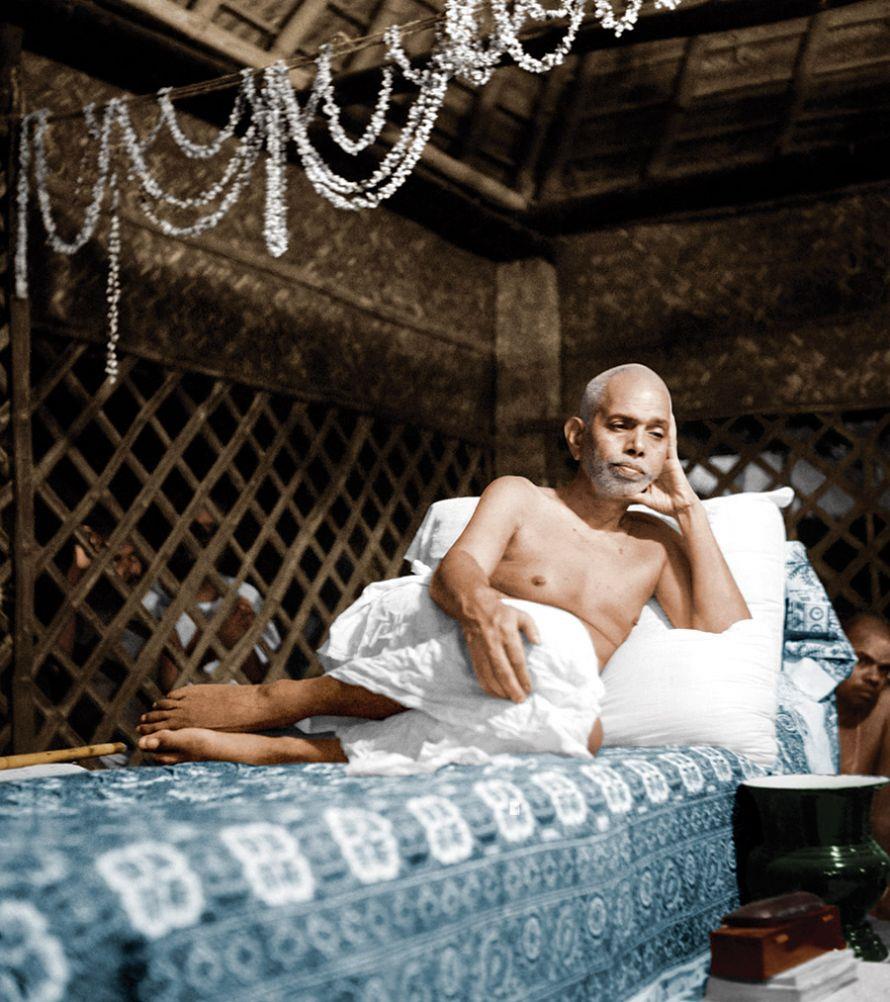 full-page colour photo of Sri Bhagavan reclining on his couch in a thatched shed hung with garlands